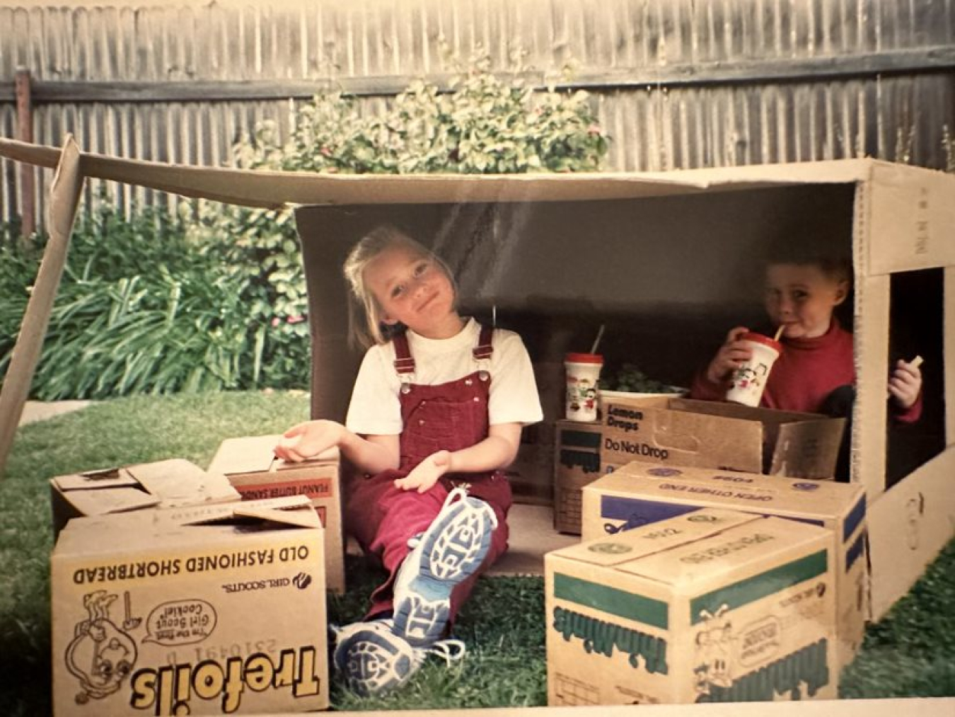 Maxine Hillman, as a child, sitting in a large cardboard box with another child, surrounded by smaller cardboard boxes