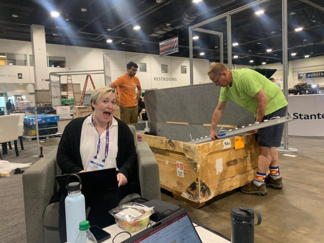 Maxine Hillman sits in a large room with a laptop on her lap while two people unload a large crate behind her