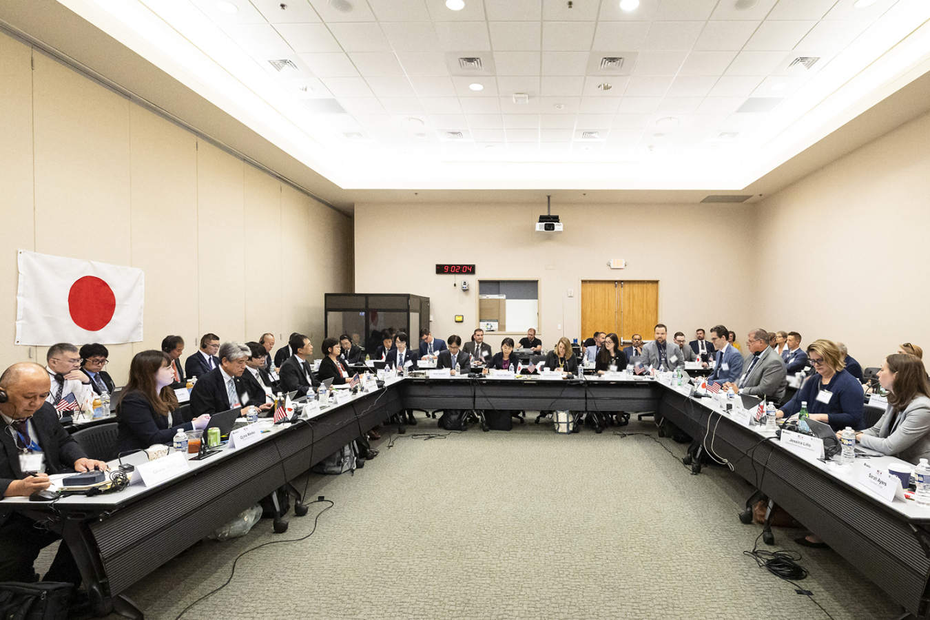 A group of people seated around a large U-shaped conference table. One side has a Japanese flag on the wall behind it.