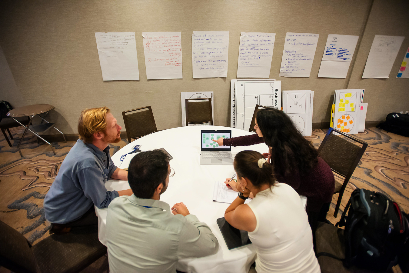 Four people sit around a table looking at a laptop in front of a wall with big sticky posters on it.