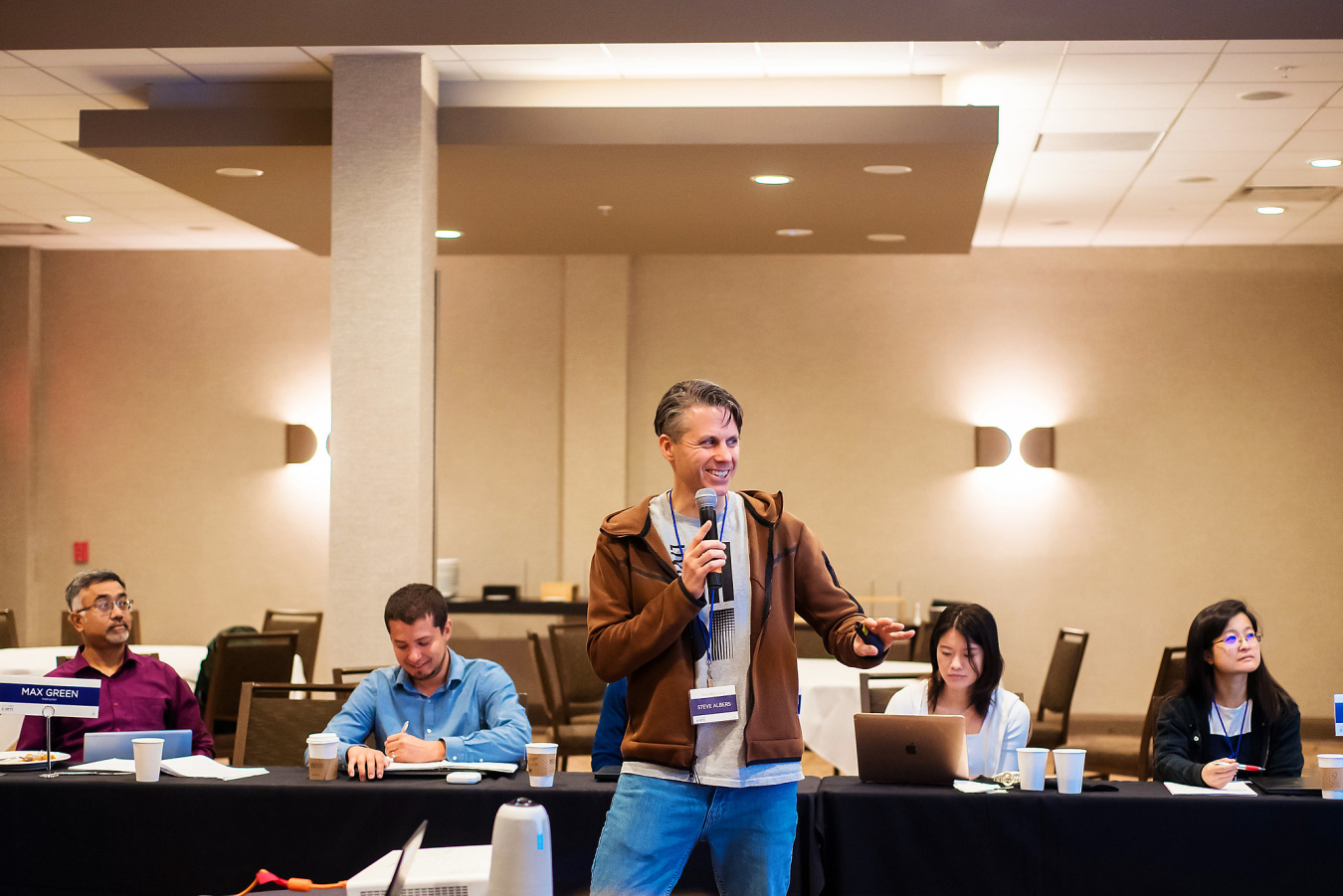 A man stands with a microphone talking to a meeting room of people.