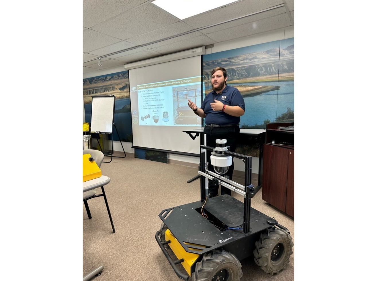 Brendon Cintas leading a slideshow presentation. A robot on four wheels sits in the floor in front of him