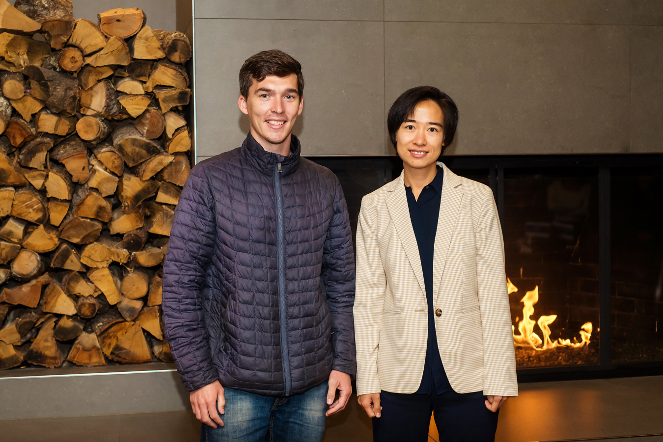 Two people pose and smile for the camera in front of an indoor fireplace.