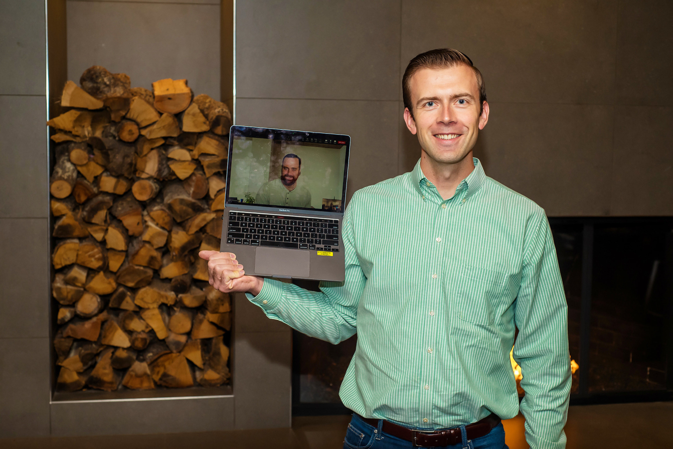 One man poses and smiles for the camera while holding a laptop showing another man on the screen.