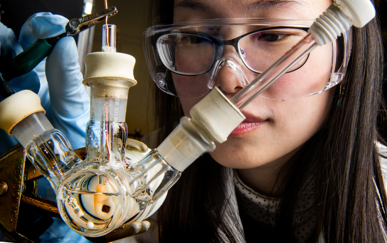 Woman with safety goggles closely examines a glass laboratory contraption that she holds with a gloved hand. 