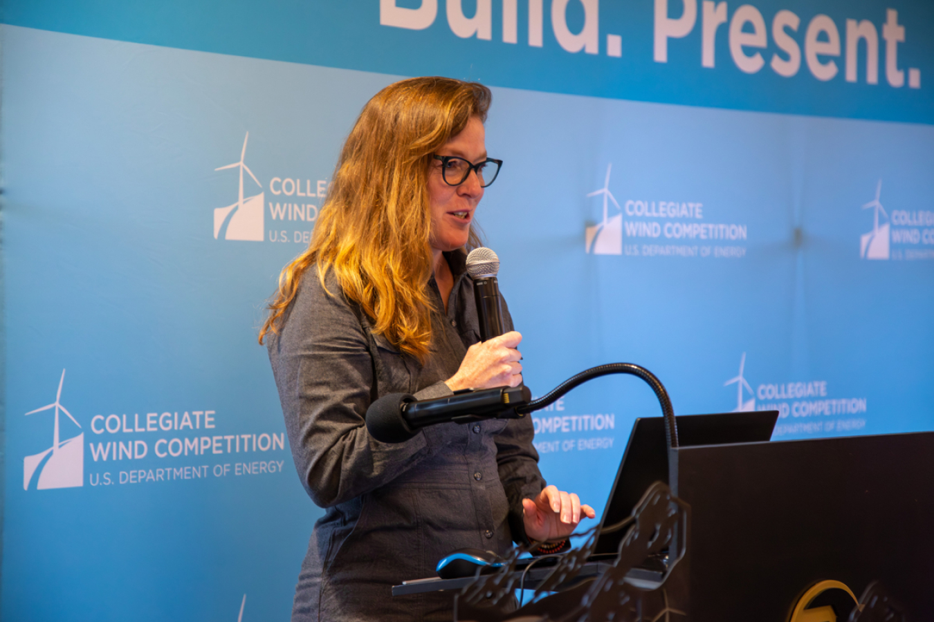 A woman standing at a podium and speaking into a microphone in front of a backdrop showing the logo for the U.S. Department of Energy Collegiate Wind Competition. 