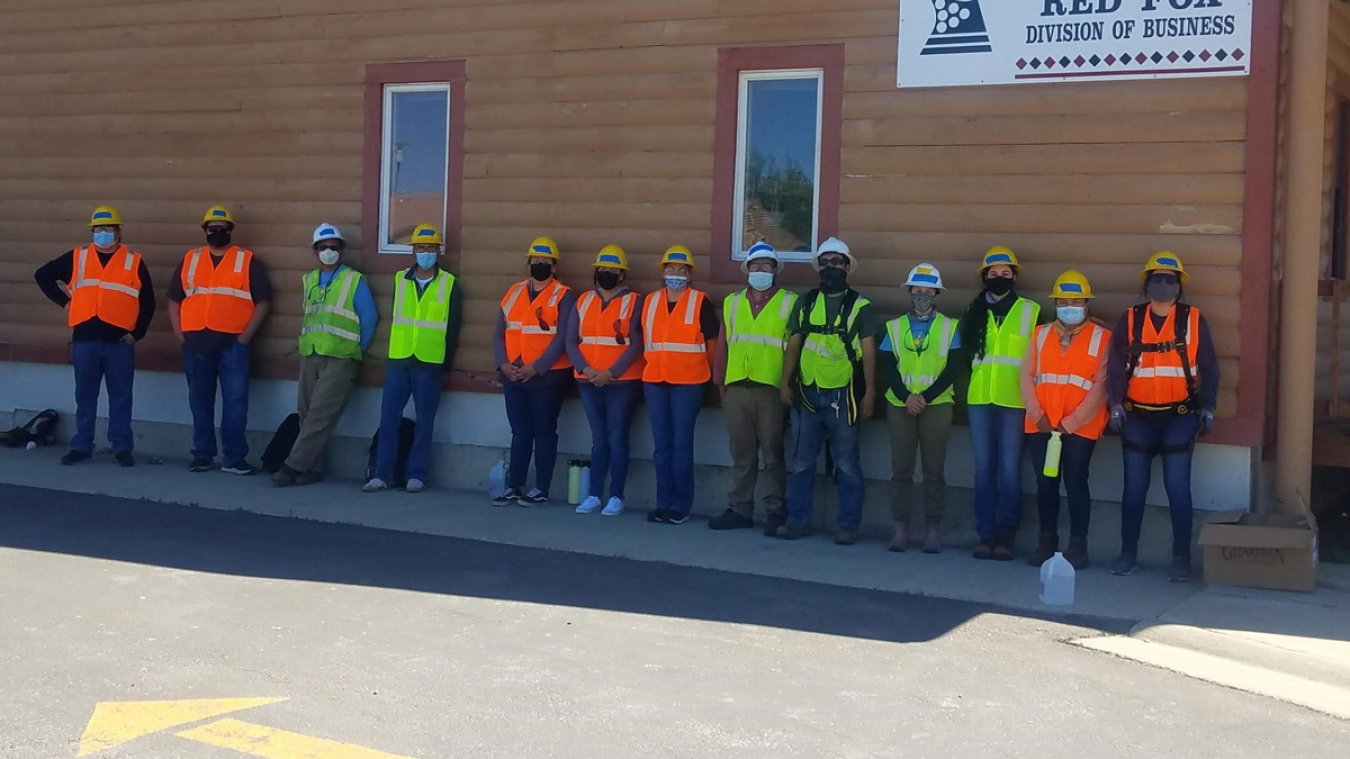 13 students wearing construction hats and vest stand in the shade along a building.