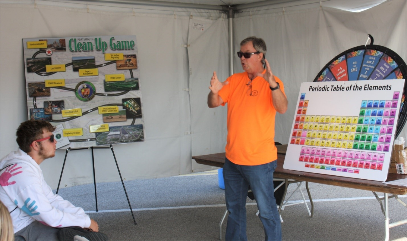 A man stands beside a poster of the periodic table and gives a presentation to students