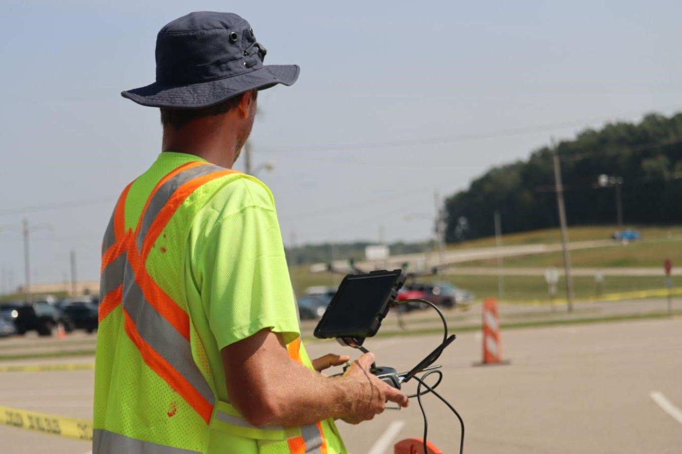 A man in a neon yellow safety shirt controls a drone with an iPad and controller