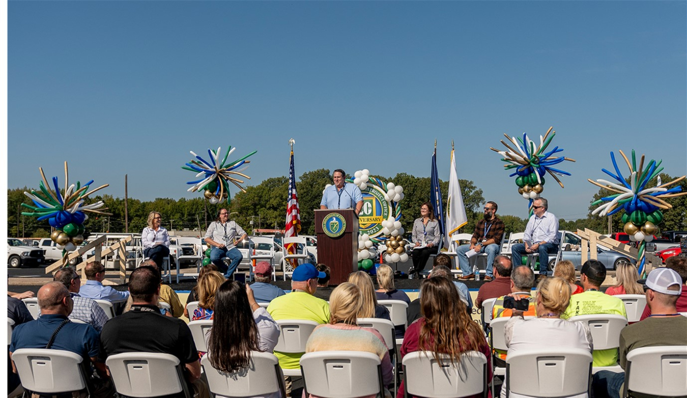 Speaker at podium addresses crowd at celebration event outside.