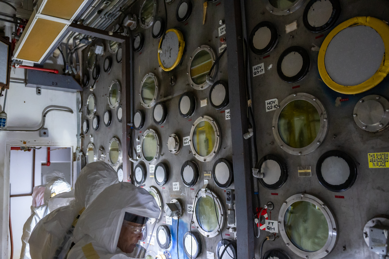 Employees in hazmat suits inspect inside a room with lots of meters on the wall