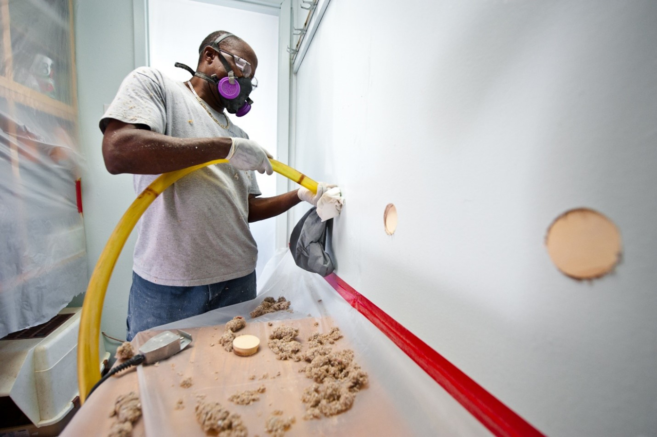 A person wearing a respirator directs a hose into a wall.