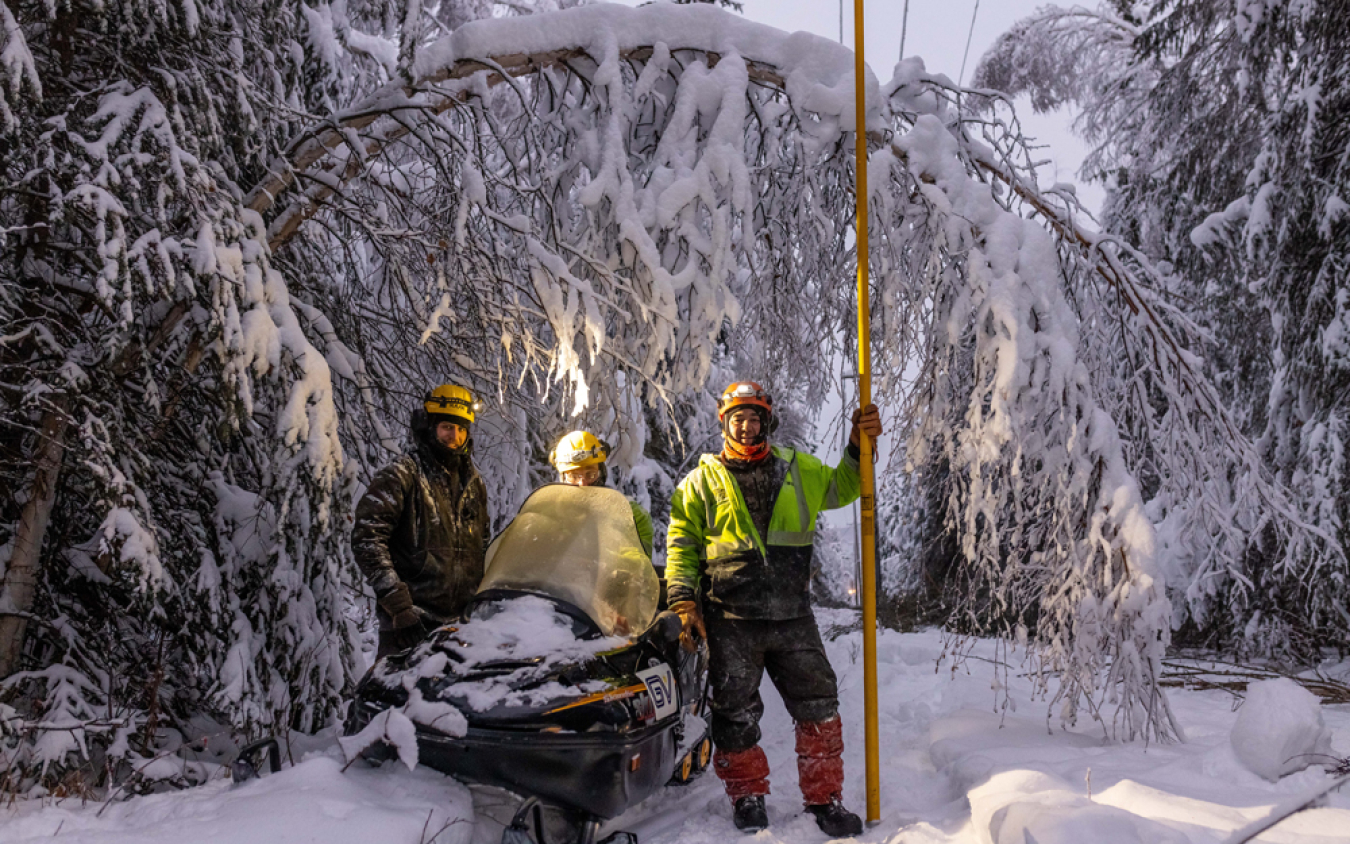 Three utility workers stand alongside equipment and a snowmobile in a snow-covered landscape. A tree branch bends under the weight of heavy snow behind them.