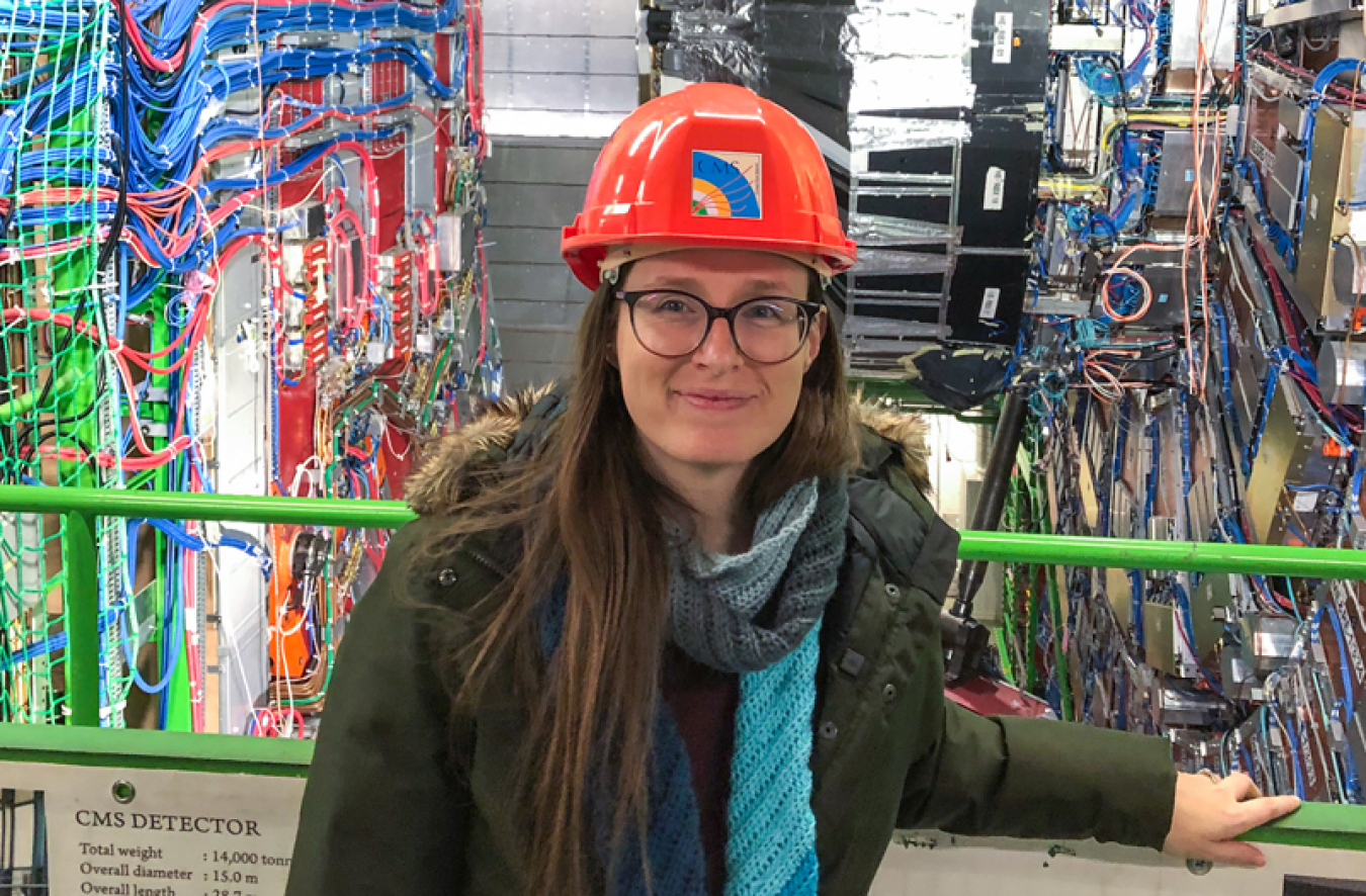 Nadja Strobbe (a white woman in a green jacket and red hard hat) standing in front of the detector in its open position showing silver-colored detector elements and red and blue wires.   