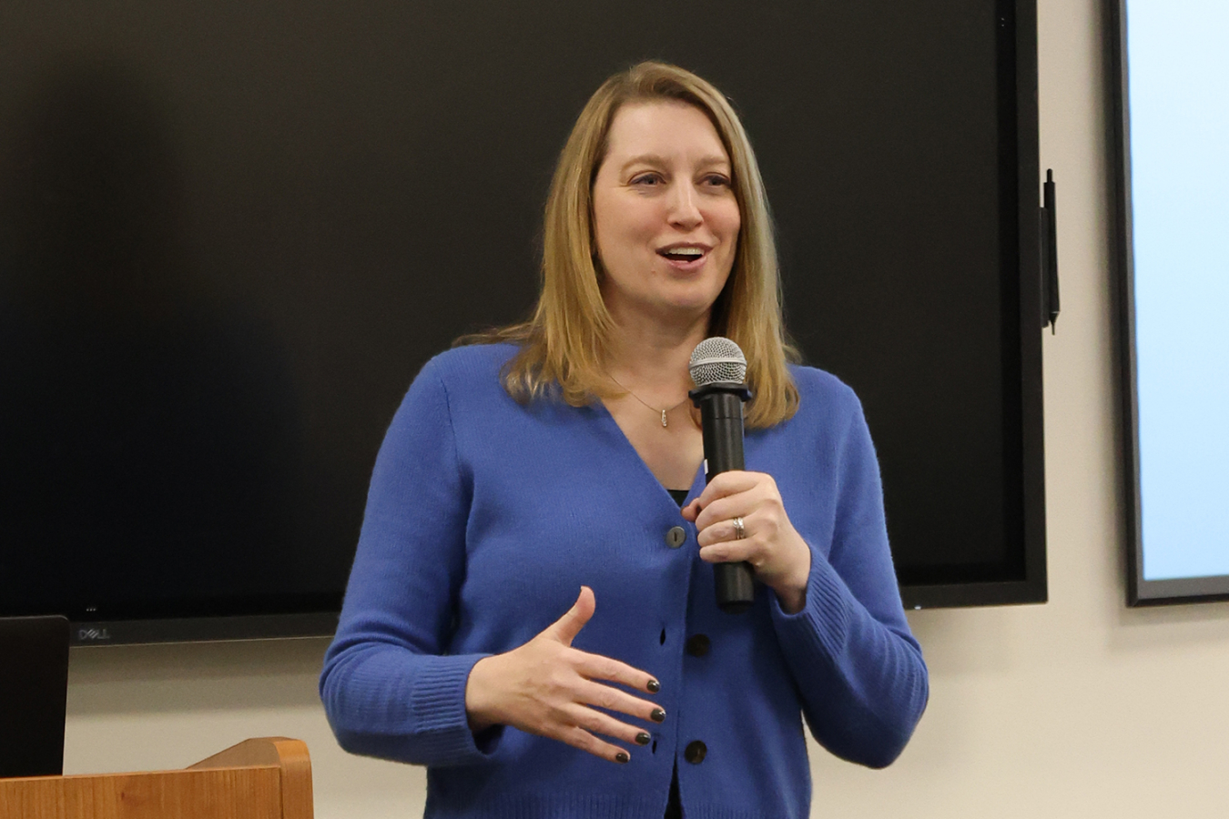 A woman dressed in a sweater holds a microphone and speaks to a group in a conference room to a group.