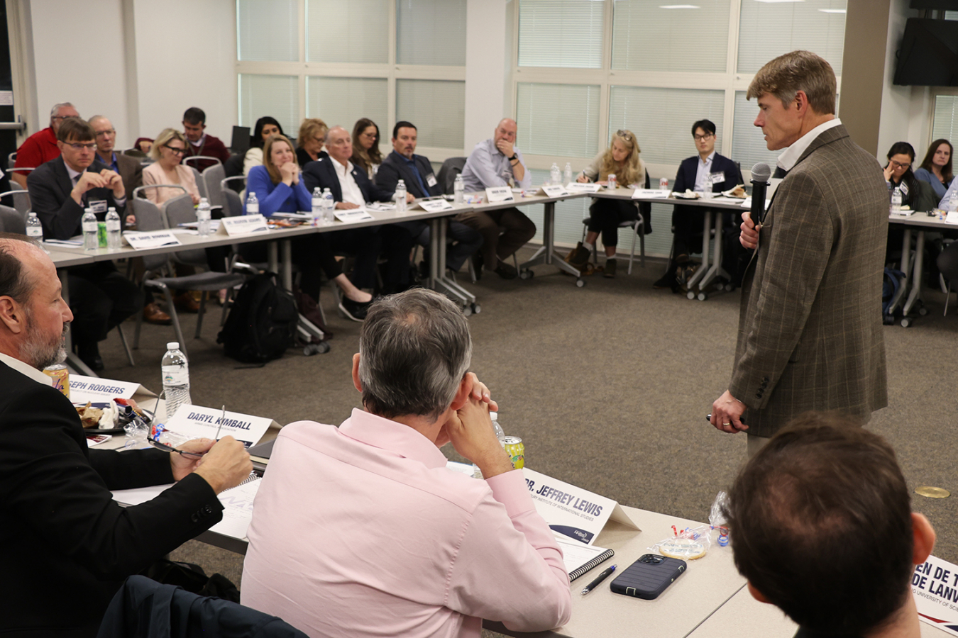 A man in a blazer talks to a group of people in a large conference room.