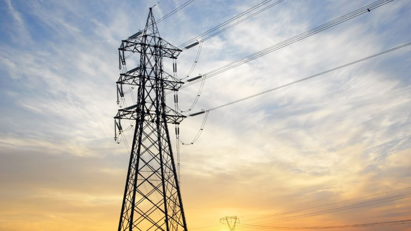 A large transmission tower and power lines appear in the foreground of the photo. In the background, the sun is setting in shades of yellow and gold against a cloudy light blue sky.