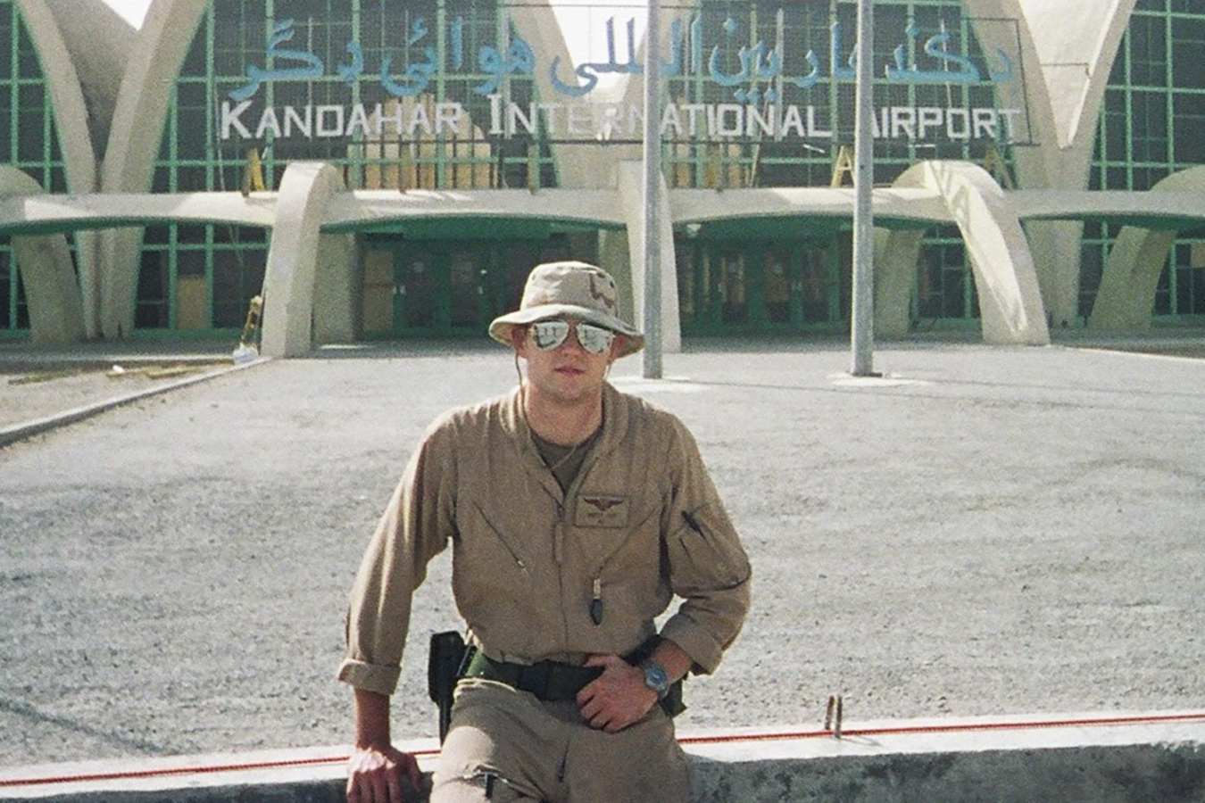 A young man in sunglasses poses in front of the Kandahar Airport sign.