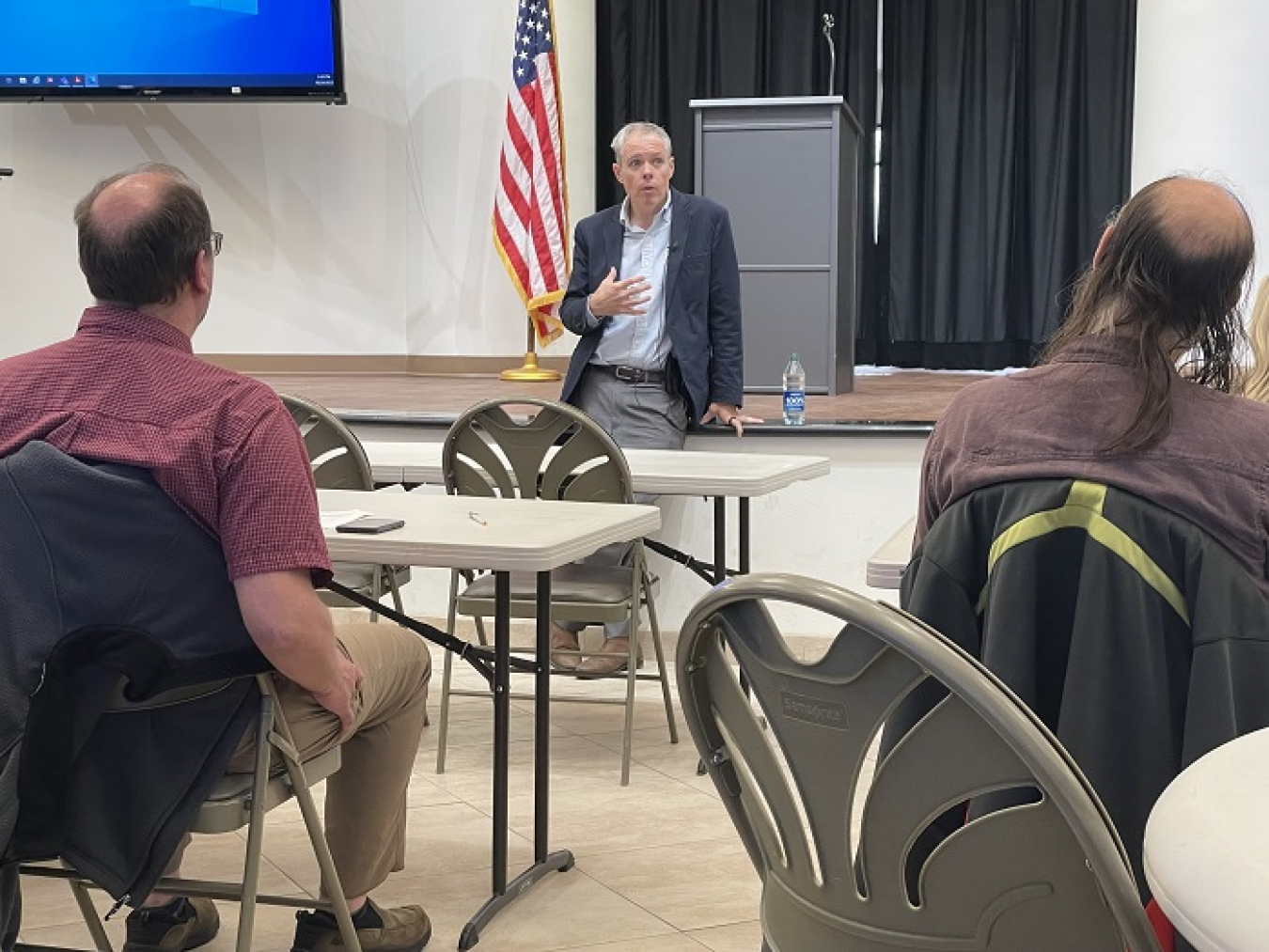 A man stands and the front of a room and speaks to people sitting at tables