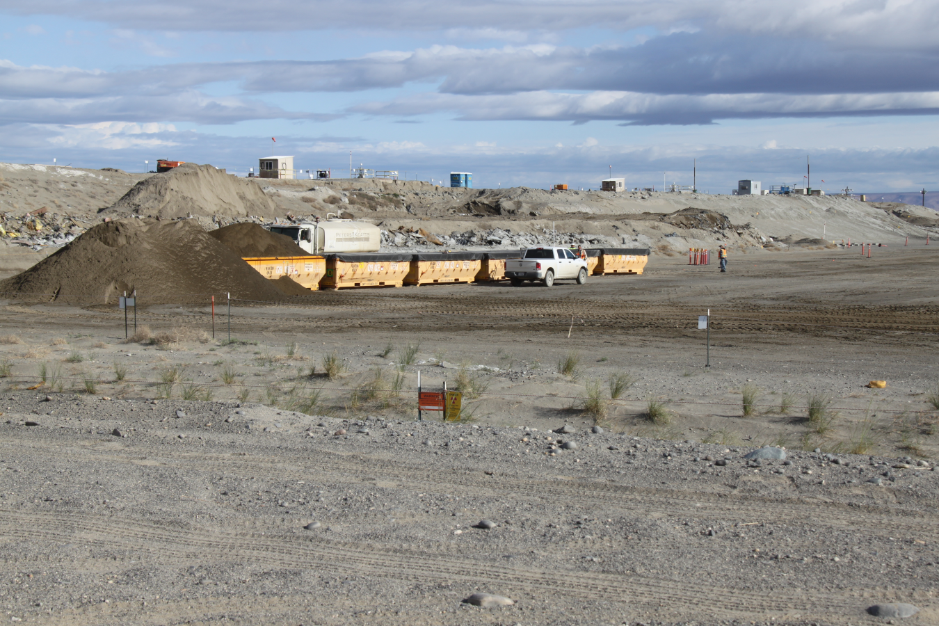 Six large yellow bins sit in the middle of a dirt lot with a white truck parked in front of them 