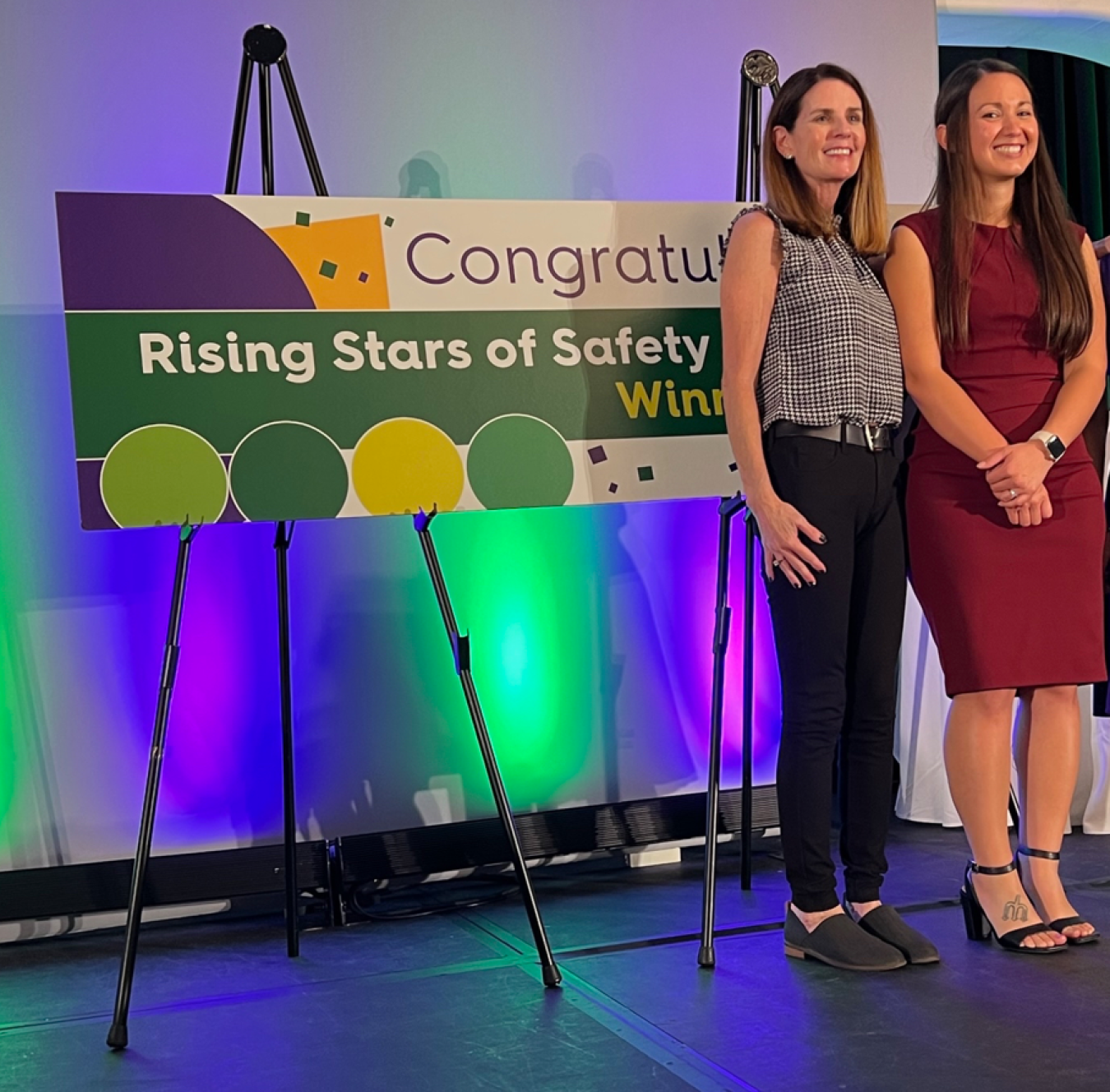 Two women pose in front of Congratulations Rising Stars of Safety poster display.