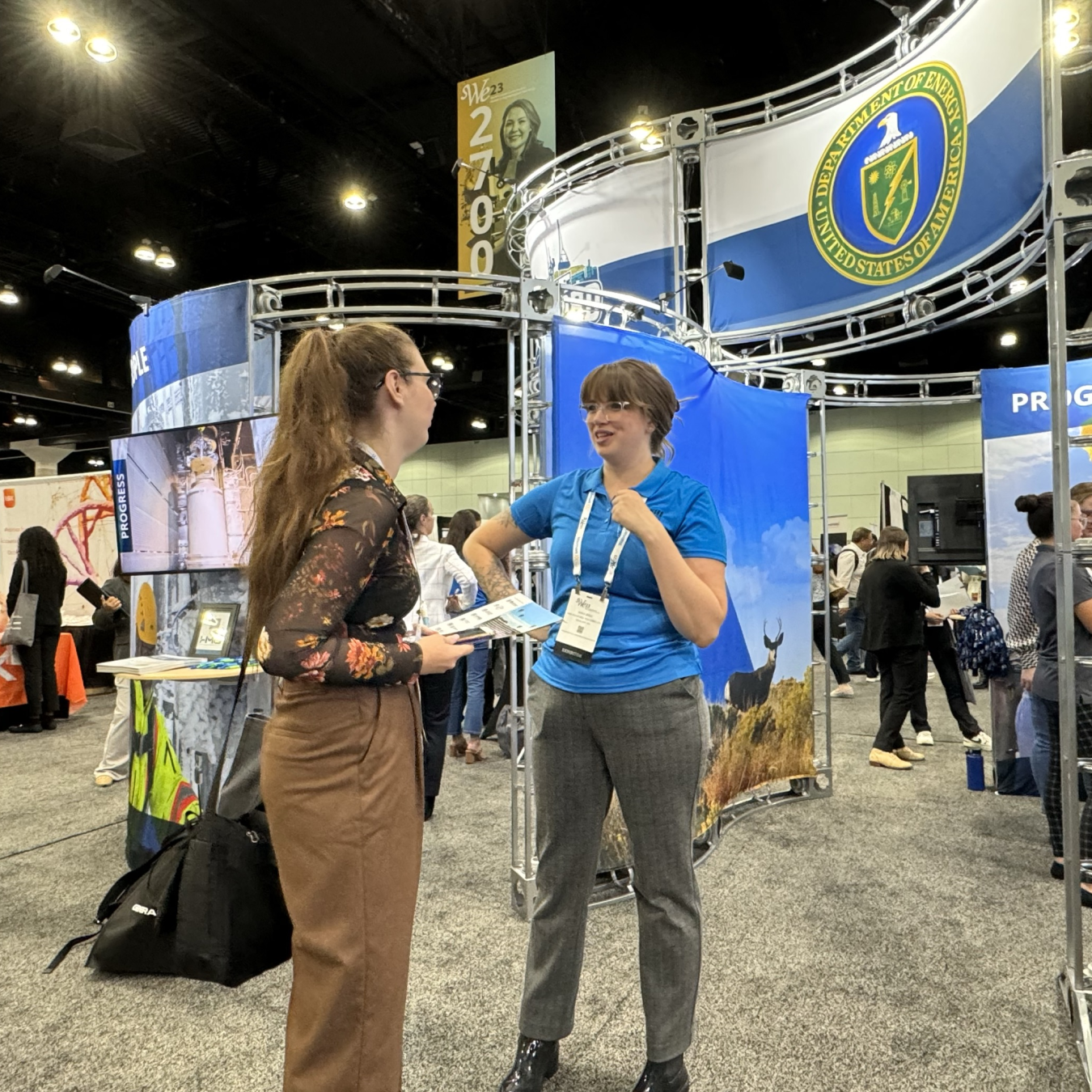 A person in a blue shirt stands beside a person in a black shirt and speaks to them 