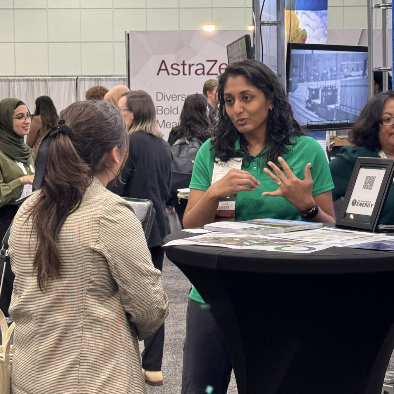 Woman in a green shirt stands at a table and speaks to a person with their back turned