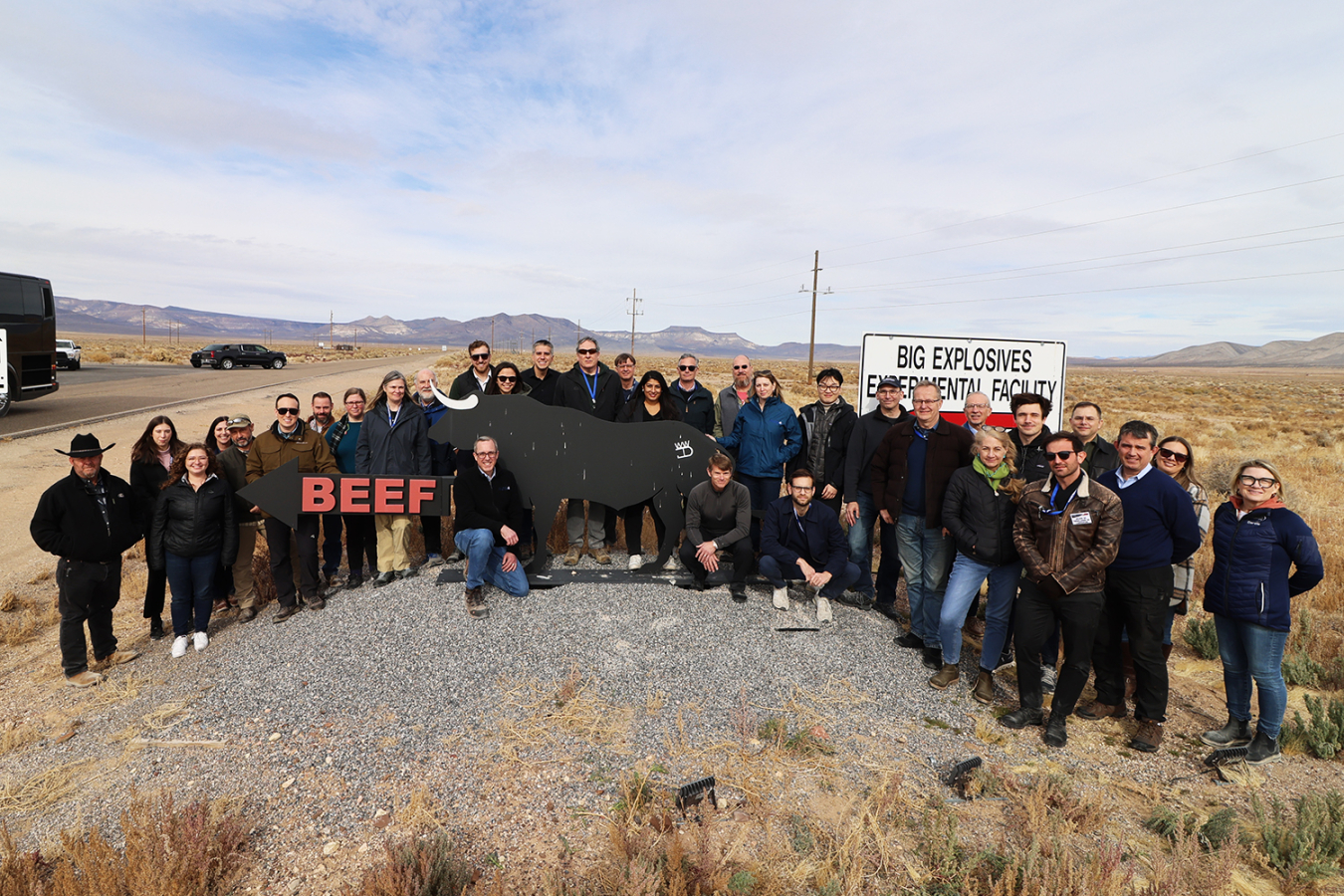 A large group of people stands outside near a sign that looks like a steer and the words Big Explosives Experimental Facility.