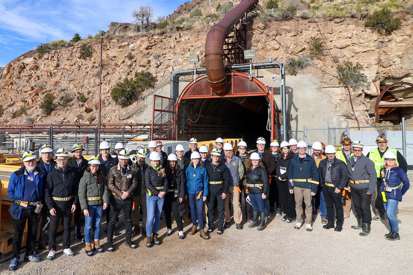 A large group of people stands outside in front of a mine entrance. To the left of them is a mine railway.