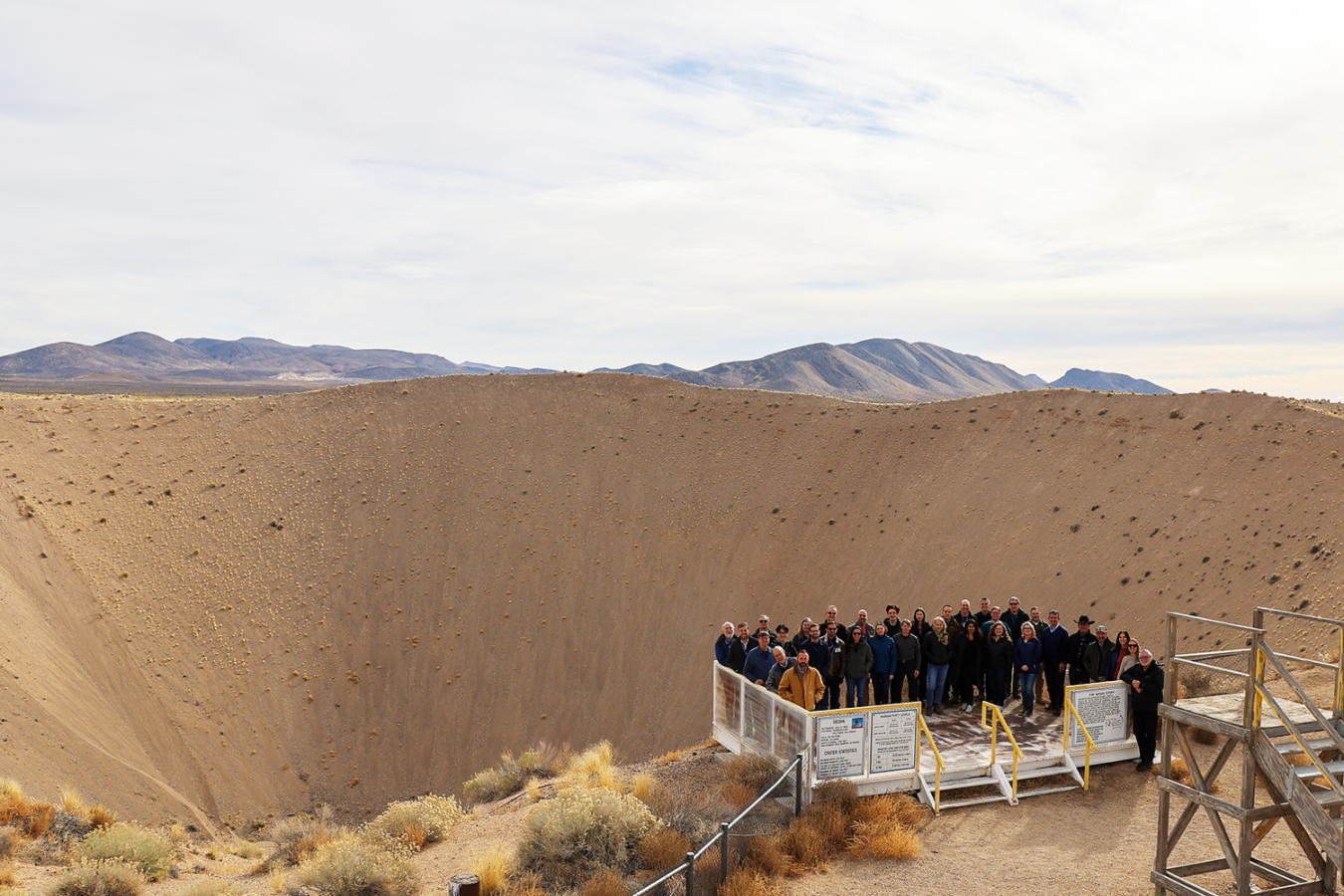 A large group of people stands outside on a metal stand on the precipice of a large crater.