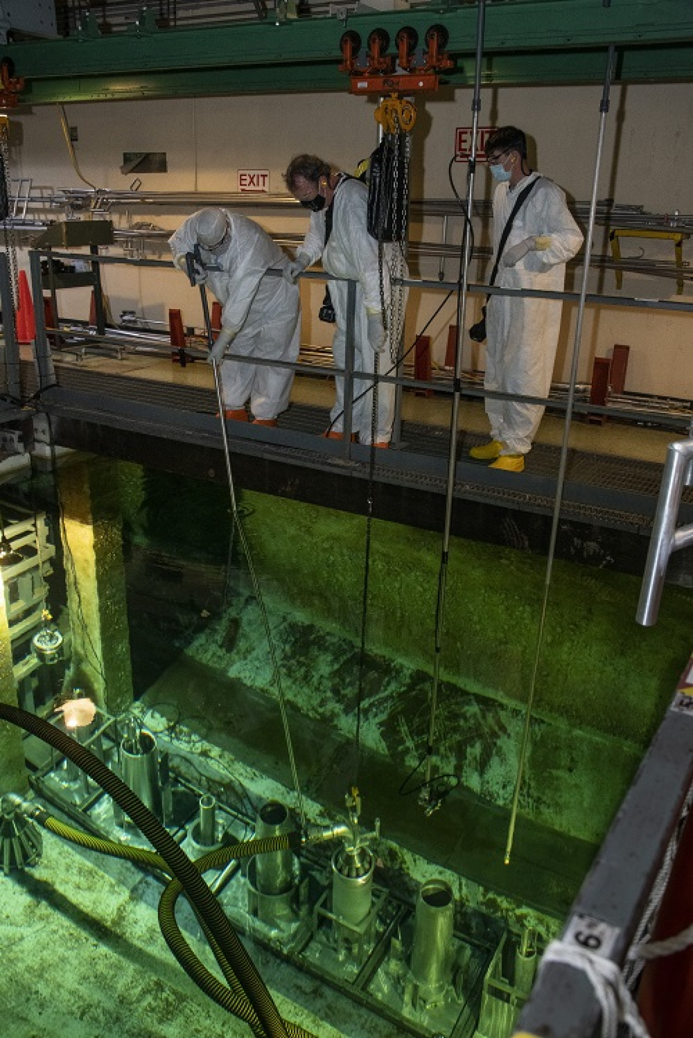 Inside of a dark basin. 3 workers in white suits stand on a walkway and look over