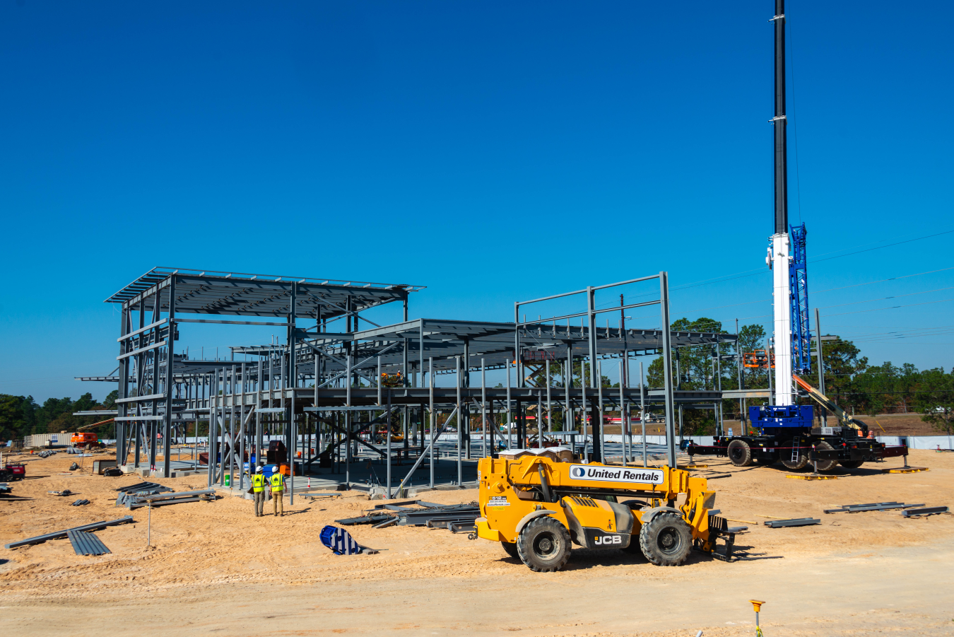 Steel building structure with two workers in safety vests and two construction vehicles