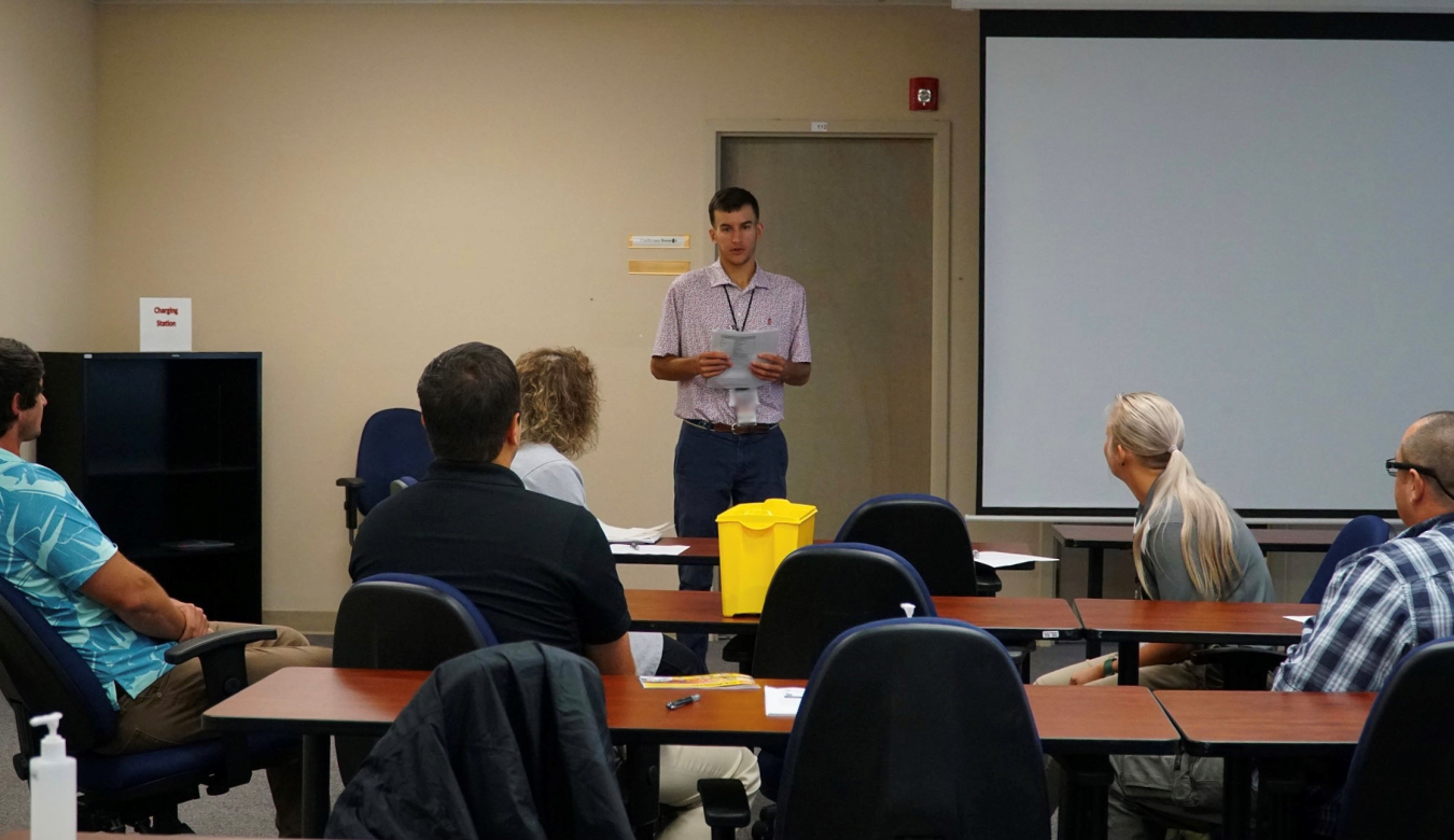 A man stands at the front of a classroom speaking to adults sitting at tables