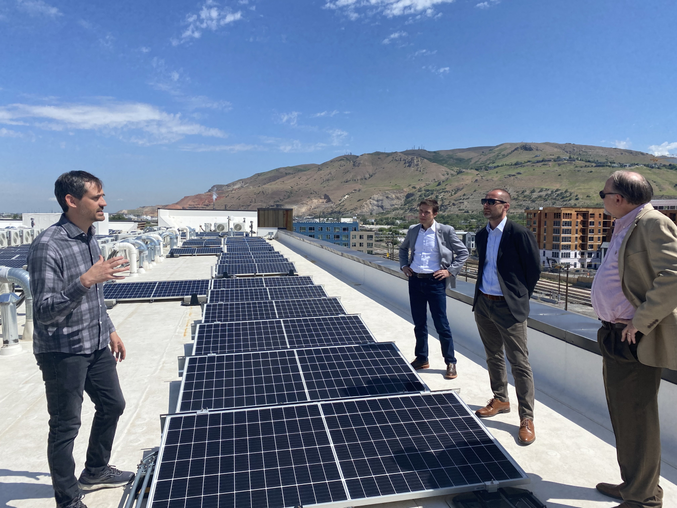 Four men standing next to a series of solar panels on a rooftop, with mountains showing in the background.