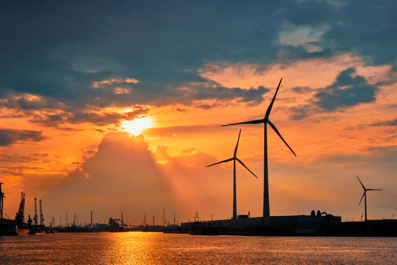 The sun sets on wind turbines near a port. 