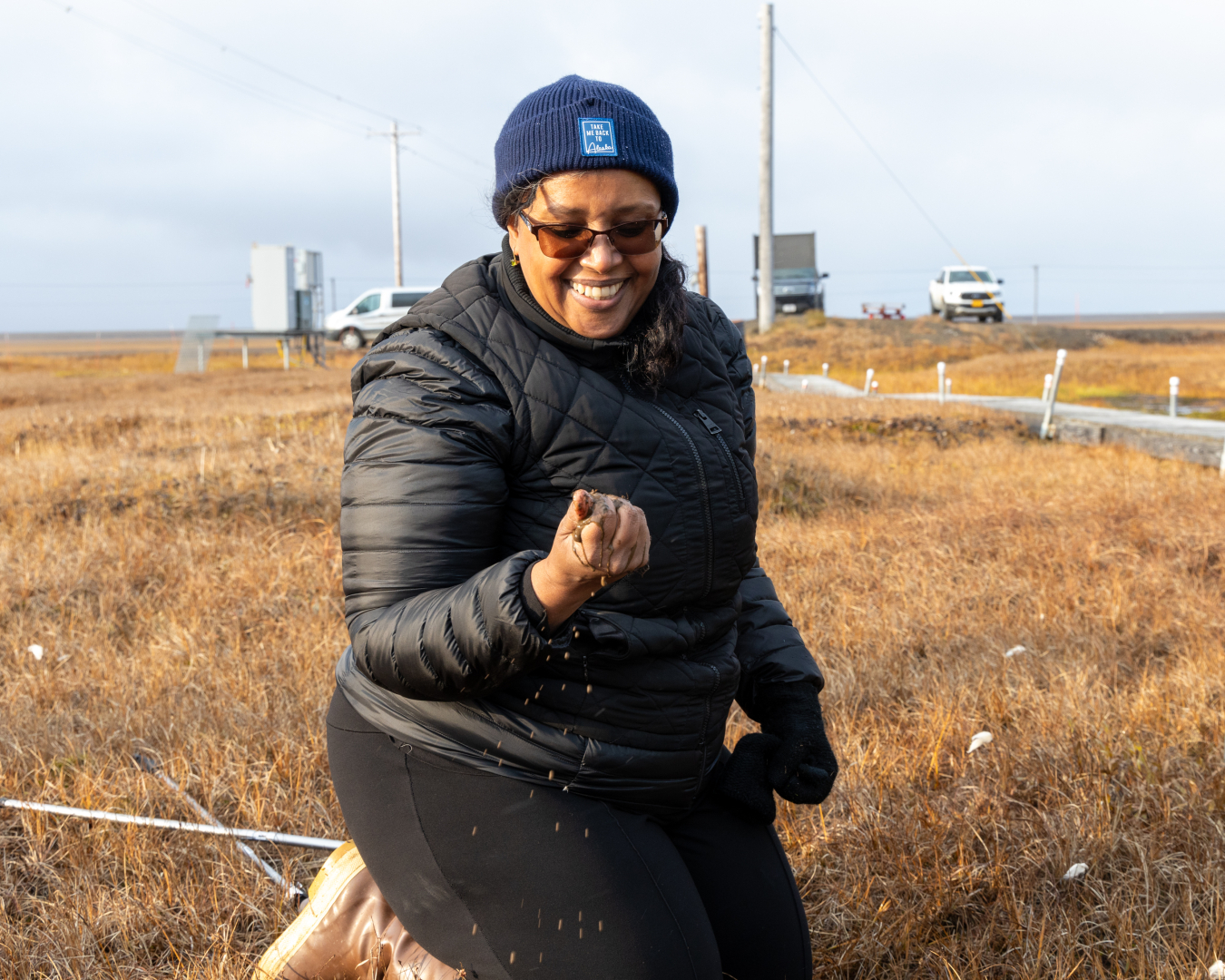 Asmeret Asefaw Berhe (a Black woman in a puffy black coat) holding a handful of dirt, standing in the tundra with dried grass behind her