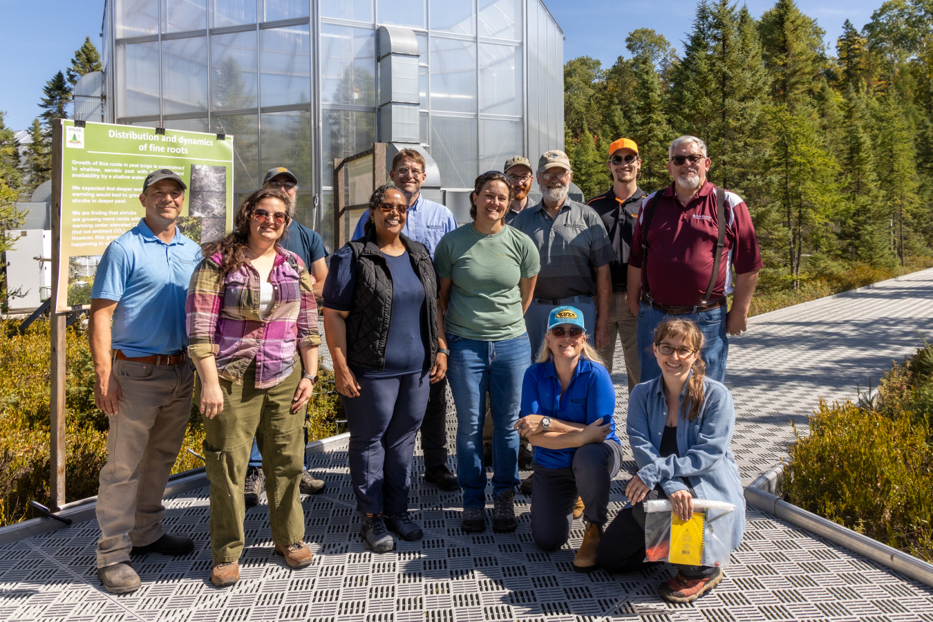 A group of people in outdoors clothing standing on a metal walkway in front of a large glass greenhouse