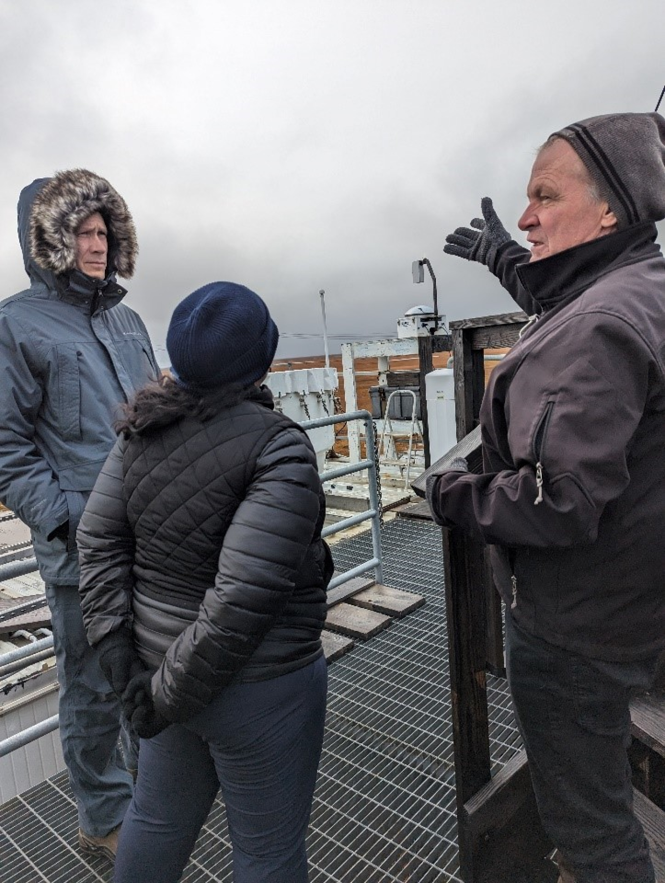 Rob Leland (a white man in a puffy winter jacket), Asmeret Asefaw Berhe (a woman in a puffy jacket and hat with her back to the camera), and Mark Ivey (a white man in a winter jacket and hat) look out on atmospheric monitoring equipment with a gray sky in the background.