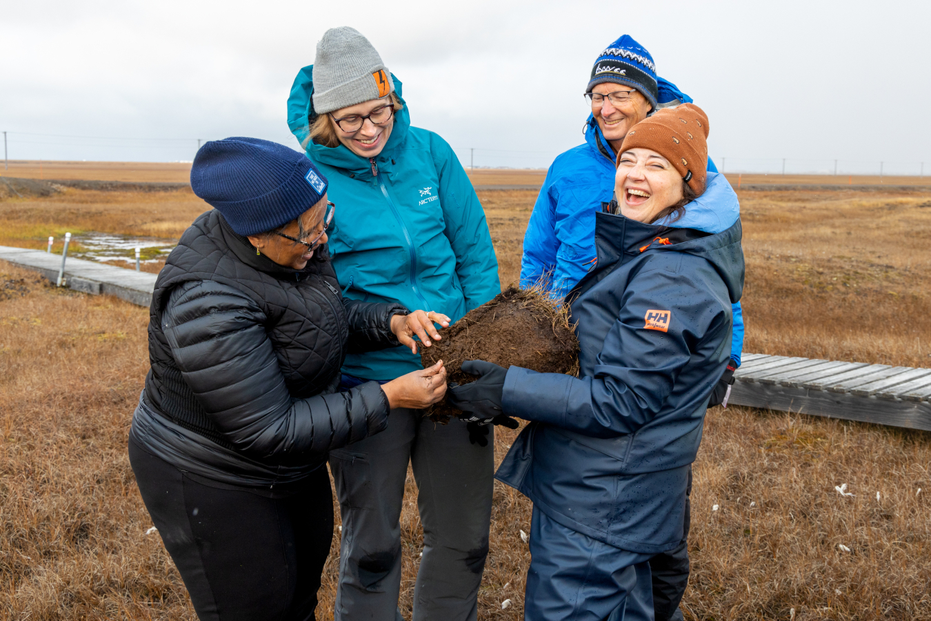 Four people in cold weather clothes standing around a chunk of soil pulled from the ground with the brown tundra in the background. 