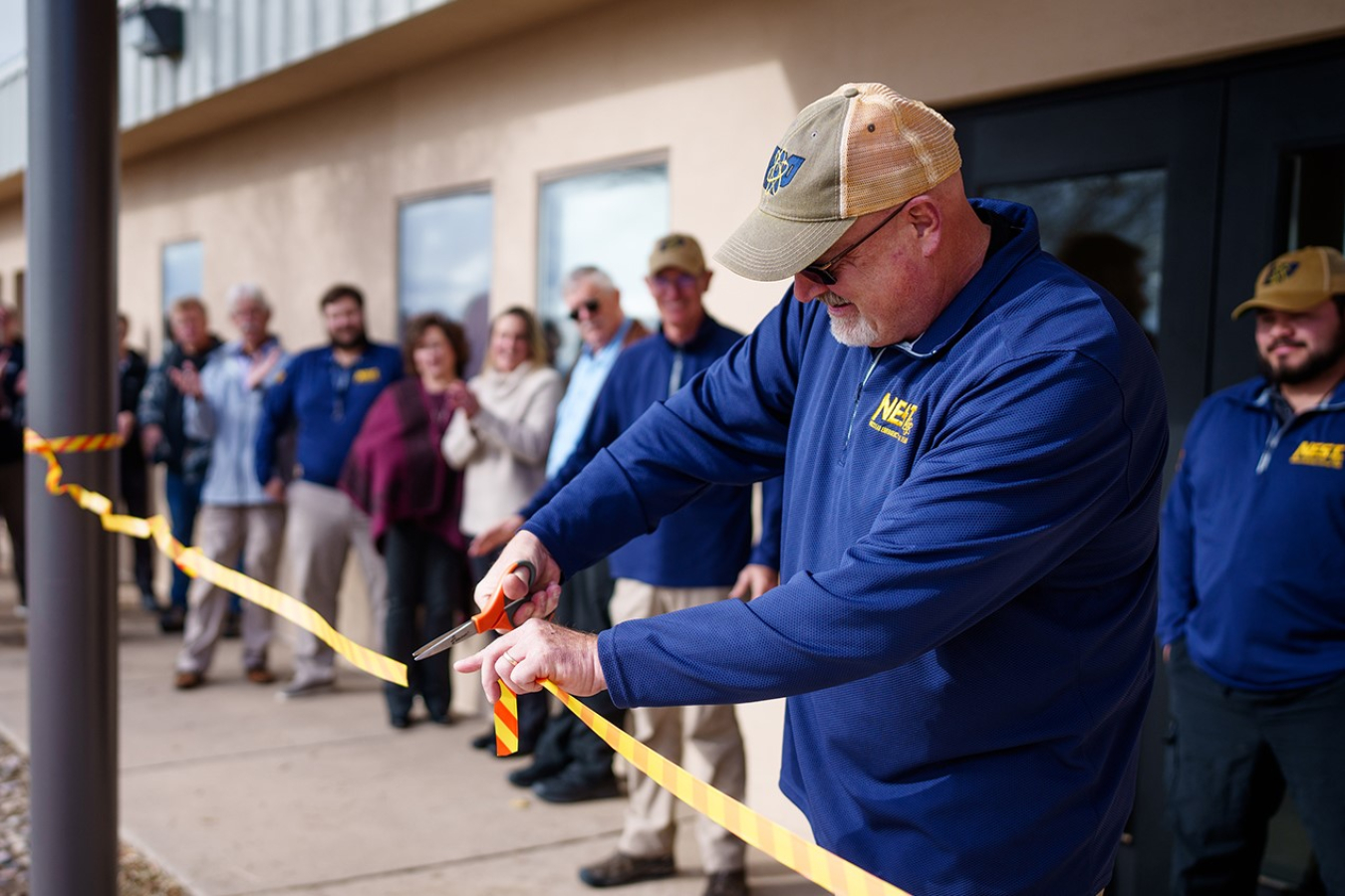 A man dressed in a NEST pullover cuts a plastic ribbon with a pair of scissors at the entrance to a building as others look on.