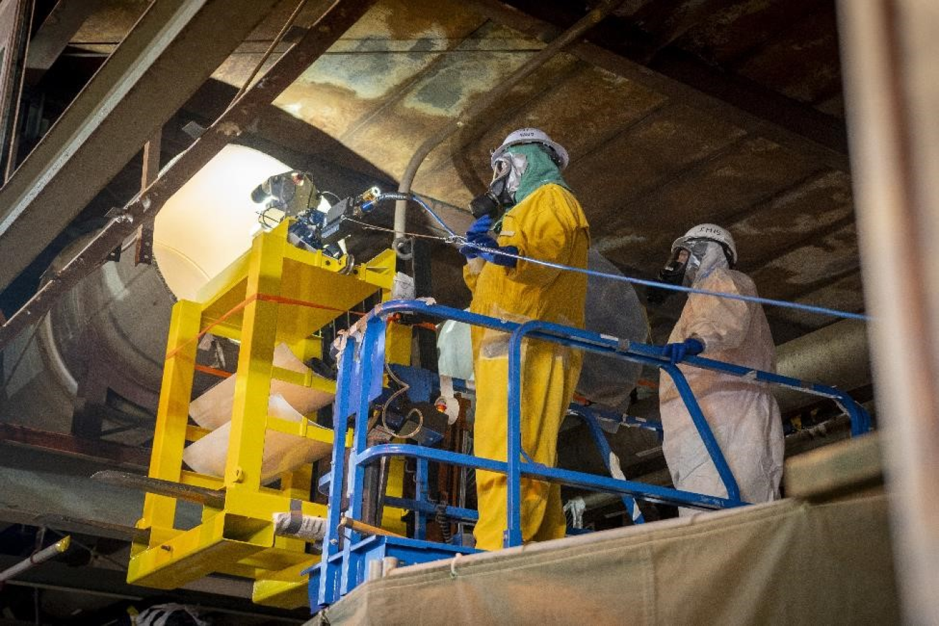 Employees in yellow and white hazmat suits lift a pipe into place