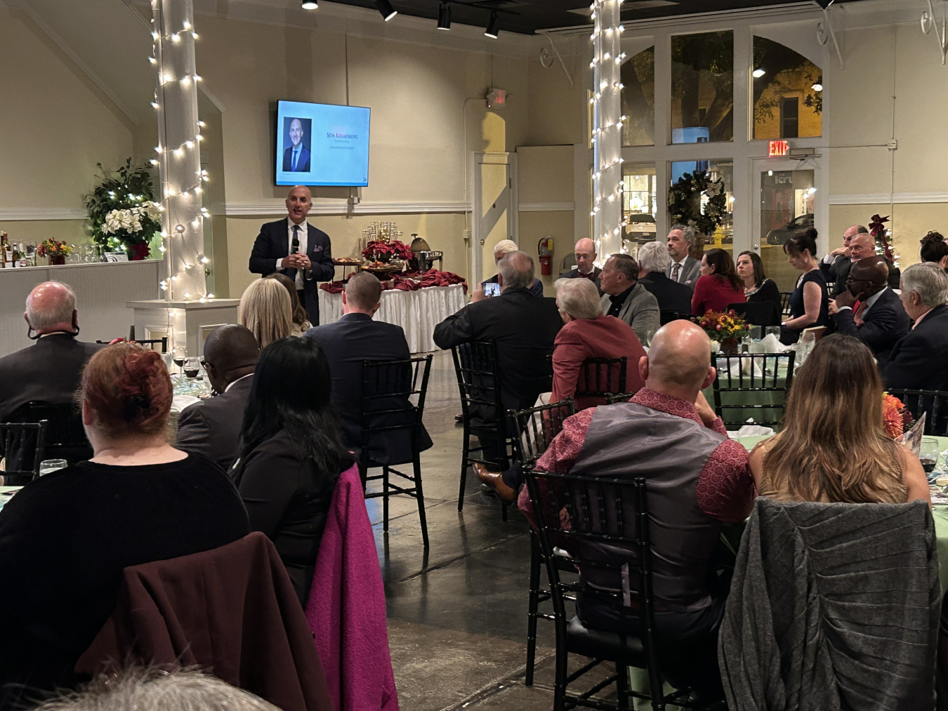 People sitting at tables warch and listen to a man at the front of the room