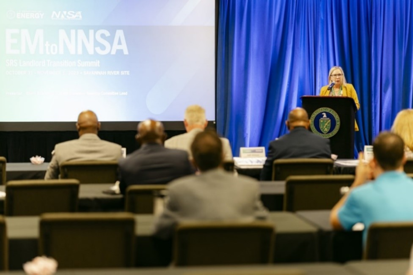 A woman stands and speaks at a podium on a stage in front of an audience