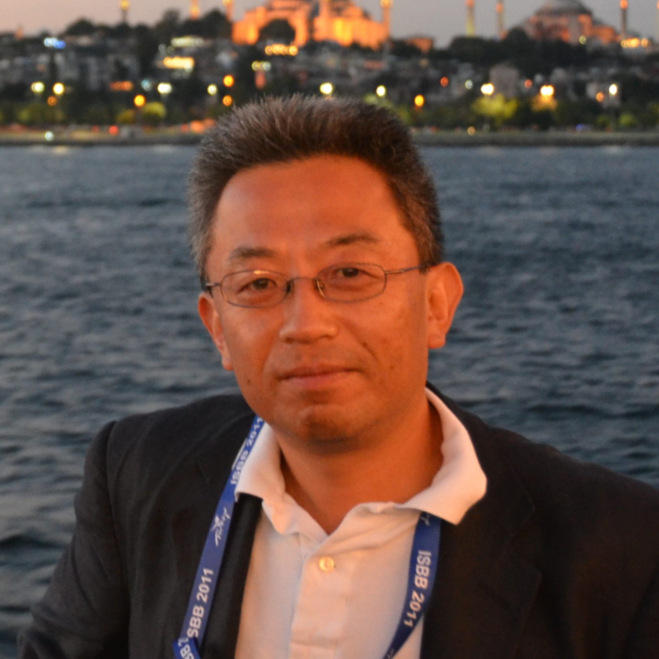 Tadashi Ogitsu, smiling, wearing glasses, a blazer, and collared suit. In the distance, there is a body of water and a line of buildings on the other shore.