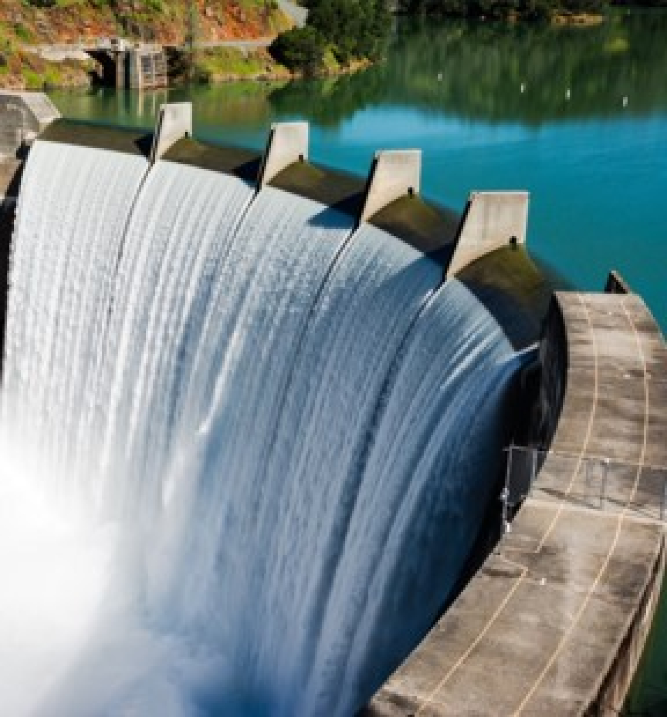 An aerial image of clear, still blue water flowing through a hydroelectric dam. White falling water and mist can be seen in the foreground.