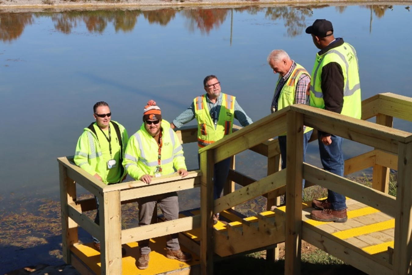 Five men in neon yellow safety jackets stand on a dock over a pond