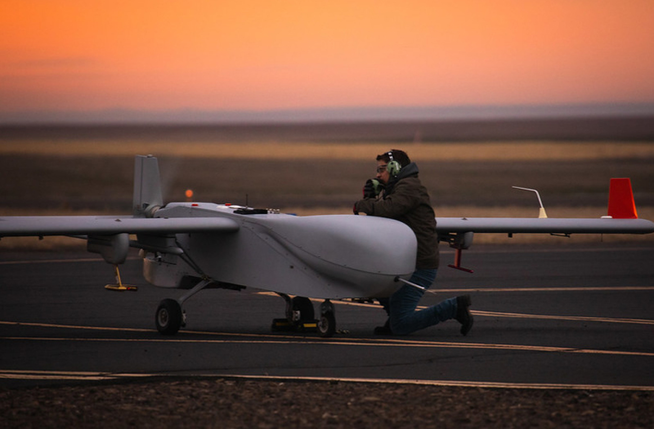 Jonathan Stratman (a man in a black puffy winter coat and jeans) kneeling next to a white aircraft with a very short, small body and large wings. They are on a runway with the orange sky of the sunrise in the background.