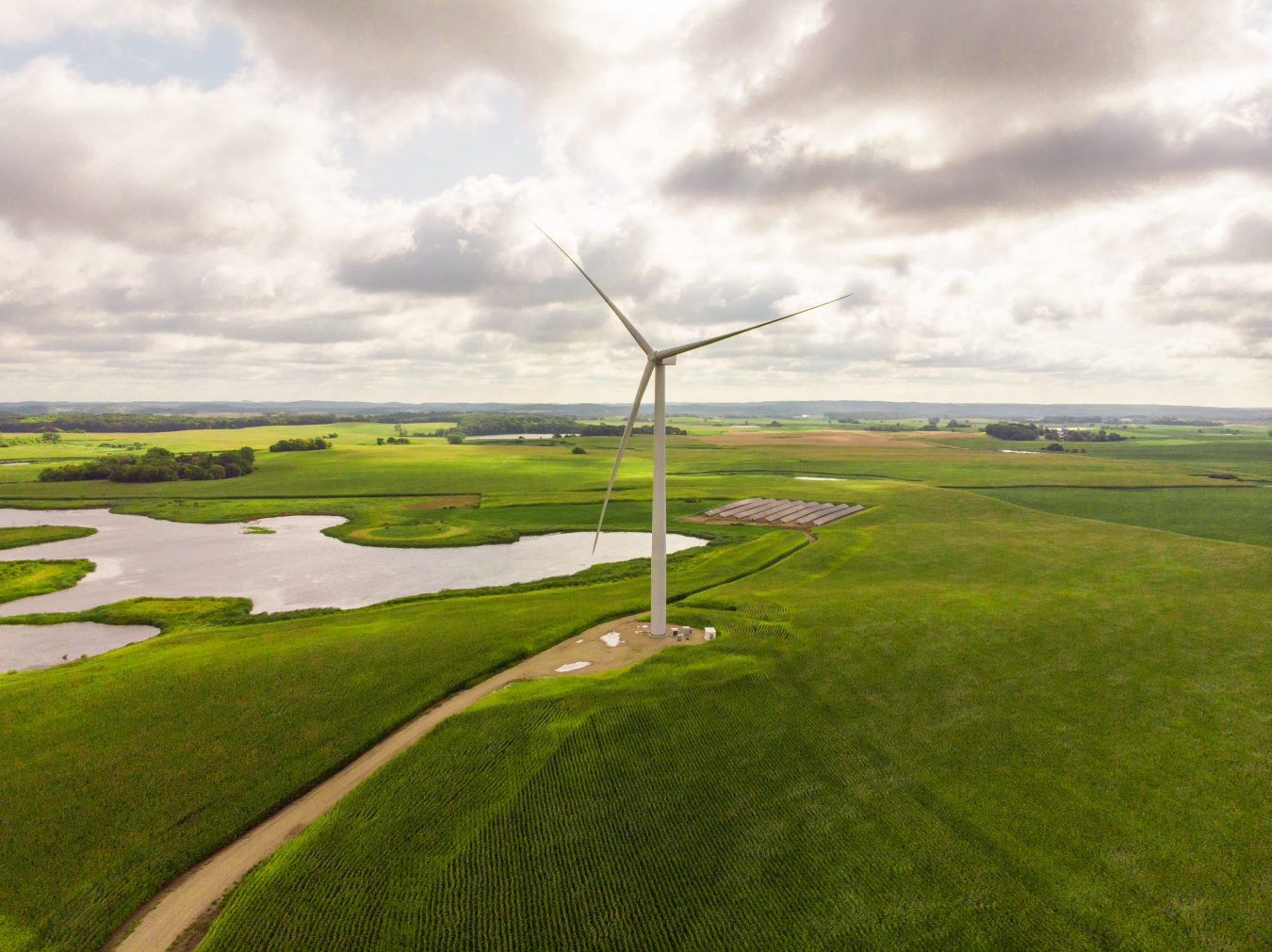 A large wind turbine on a grassy plain near a solar photovoltaic array and a lake.