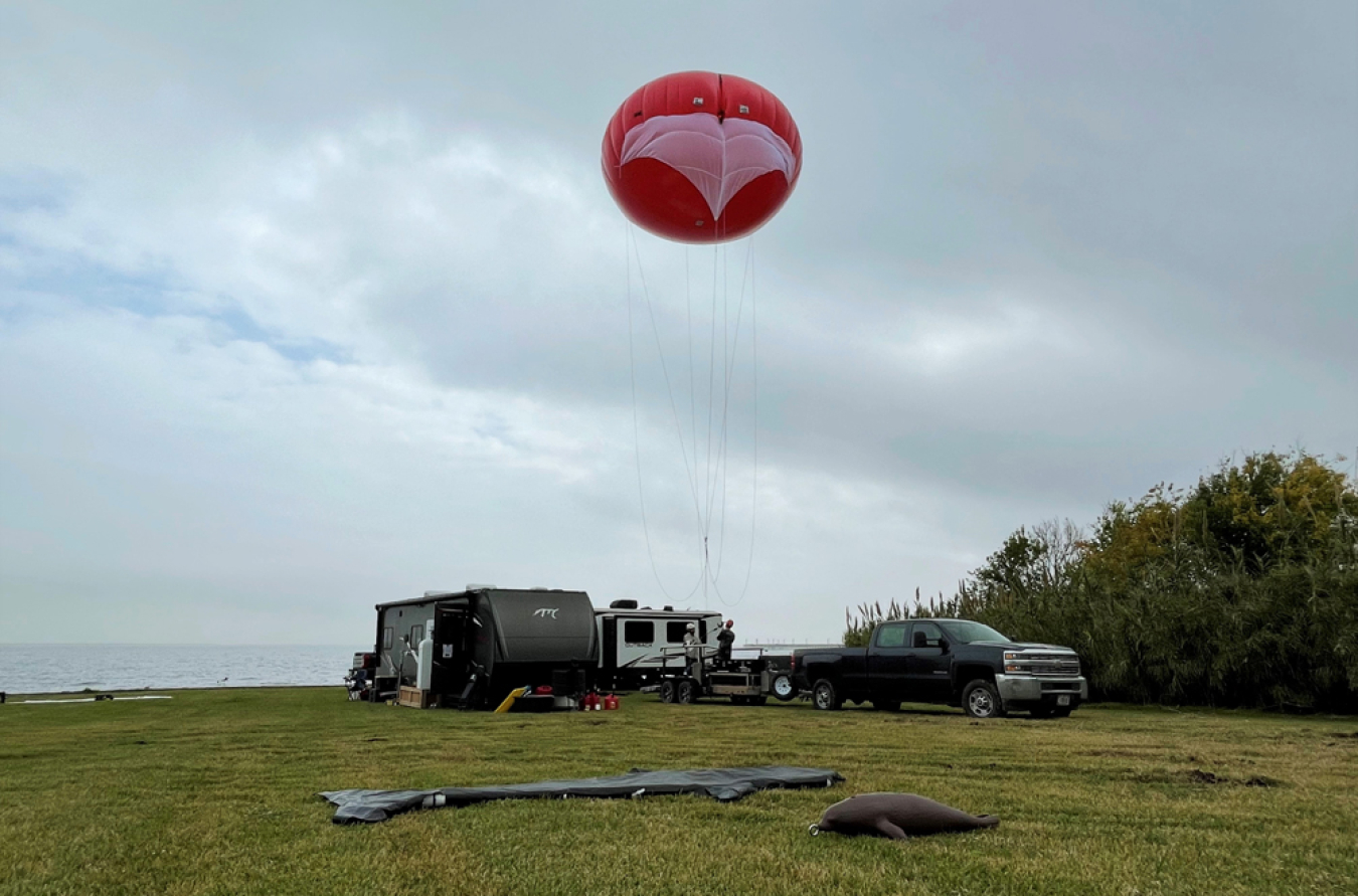 The team prepares to launch the marine wildlife detection tethered balloon to test its capabilities. Fake marine animals, made of plastic and cardboard in the shape of a seal and large fish, lay in the grass for the wildlife detection system to identify.