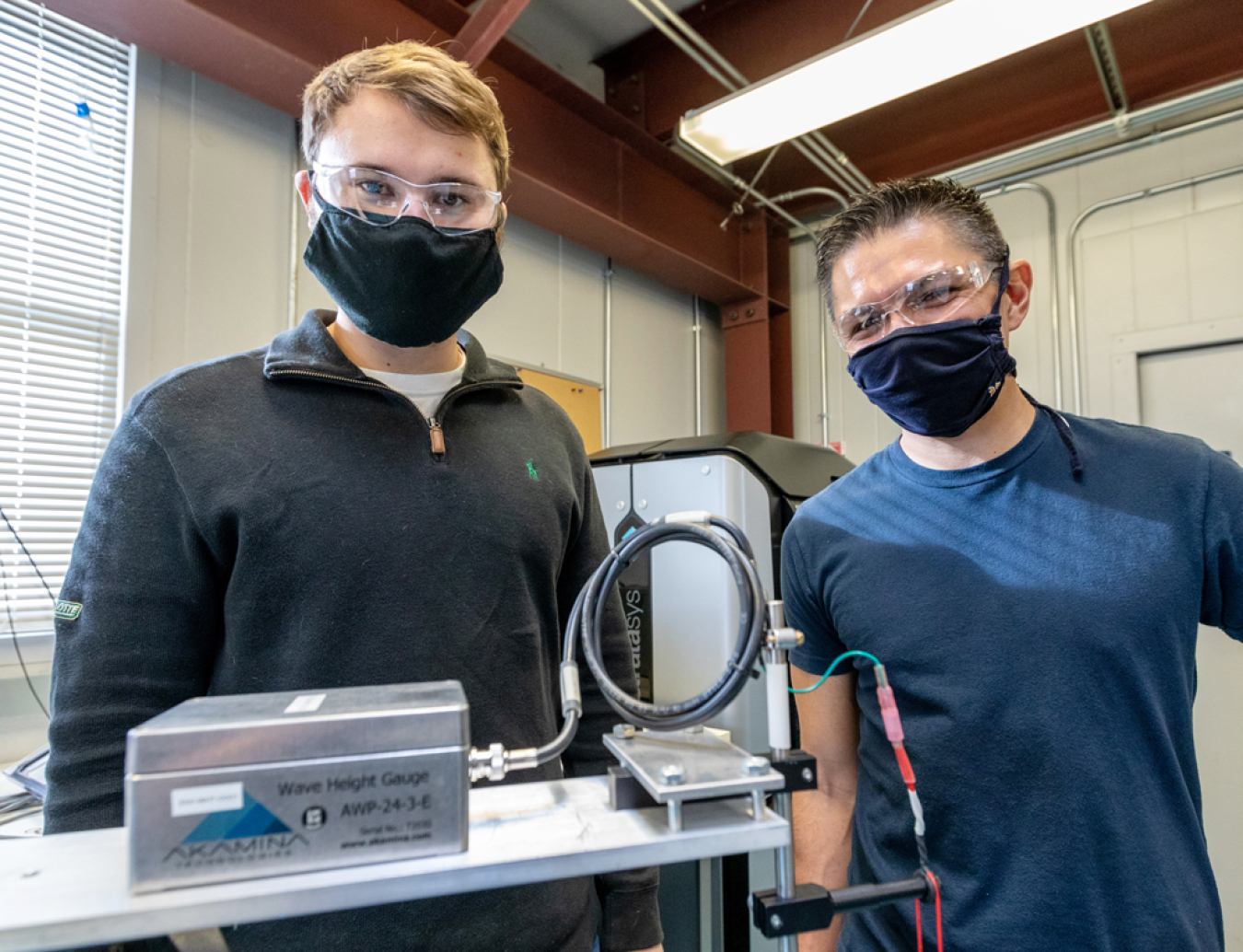 Casey Nichols and Andrew Simms wear face masks while standing next to a metal box and wiring.